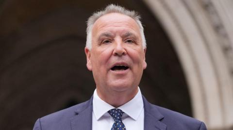 Chris Whitbread has short grey hair and is speaking in front of a cream archway at the High Court. He is wearing a navy suit jacket, white shirt and navy and yellow tie.