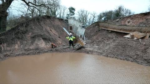 A man in a yellow hi vis jacket and white hard hat stood beside water in a hole after a canal subsided