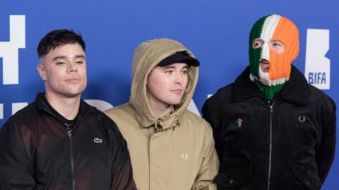 Kneecap at 2024 BIFAs - left to right Móglaí Bap in black Lacoste zip jacket, Mo Chara in a cream jacket with hood up, wearing a black snapback and pointing at camera, and DJ Próvaí in black jacket and green white and orange tricolour balaclava