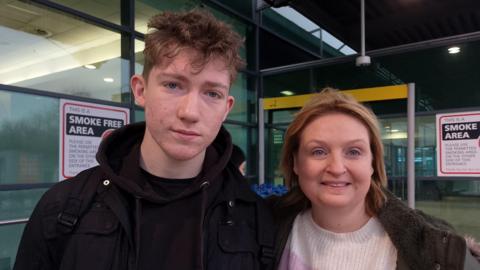 A young man, wearing a black hoodie and a black jacket, standing beside a woman wearing a cream jumper and dark green coat. 