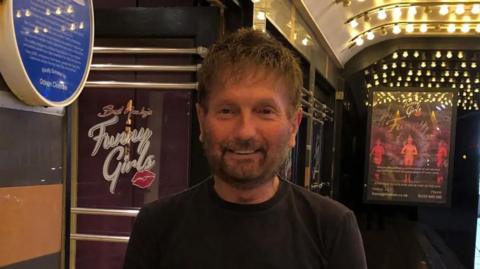 Basil Newby standing in the foyer at Funny Girls. He has brown hair and a beard and is smiling. There are posters in the background