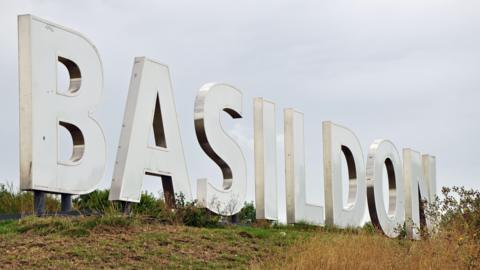 A giant white statue on a grassy bank spells out BASILDON in giant letters. It is cloudy above.