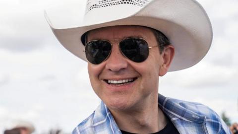 Pierre Poilievre sports a wide grin along with aviator sunglasses, a white cowboy hat, and a blue and white plaid shirt over a black T-shirt. 