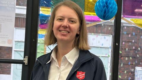 A woman with a blonde bob haircut wearing a white shirt and dark jacket which has a red badge on with The Salvation Army in white letters. She is smiling at the camera and behind her are colourful decorations and glass doors.