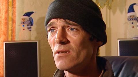 A man with a woollen hat sits in a room with Christmas decorations in the background