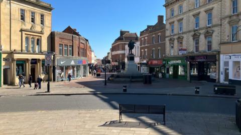 A shot of Bridgwater High Street. In the foreground is a bench draped in a shadow, while further down the road is a statue. The pedestrianised area is flanked by a number of shops. There is a blue, sunny sky.