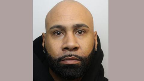 A mug shot of a man called Tyson Junior Miller wearing silver stud earrings and a black hooded jumper. He is looking at the camera and has a thick dark beard