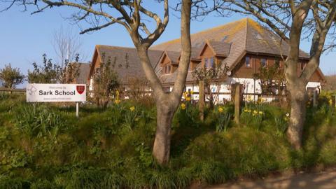 Sign reading “Welcome to Sark School” in front of a school building, surrounded by trees, grass, and yellow flowers on a sunny day.