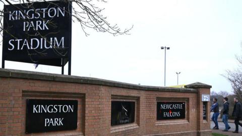 A view outside Kingston Park, home of Newcastle Red Bulls, with a Newcastle Falcons emblem on the wall as three spectators walk in   