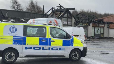 A police van sits in front of a burnt out row of shops