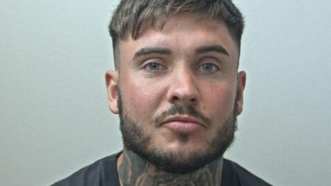 A police mugshot of Ryan Wellings, 30, who has short dark blonde hair and a short dark beard, and a tattoo covering most of the front of his neck.