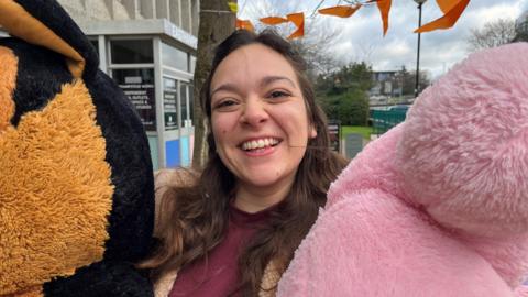 Ayesha beams as she holds two giant stuffed animals. She has long, wavy brown hair and wears a purple cotton top and fleece.