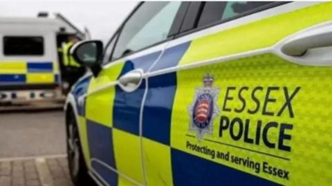 An Essex Police car parked in a bay. The car has the traditional yellow and blue panelling and an Essex Police logo on it. In the background is a white police van.