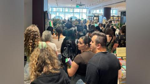 A picture inside a large bookshop at the back of a large crowd, which stretches to the far, windowed wall.