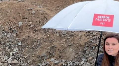 鶹Լ reporter Faye Kidd standing with an umbrella in a rock-filled crater