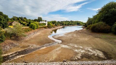 The image depicts a partially dried river or lake, with much of the riverbed exposed due to low water levels. On the left side, there are houses nestled among trees, with vegetation showing signs of dryness—some green foliage mixed with brown patches. The sky is partly cloudy with areas of blue visible, and the remaining water reflects the sky as it narrows into the distance.