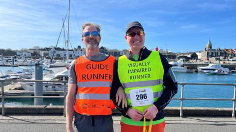 Two runners stand side by side near a marina on a sunny day. One wears an orange vest labelled “Guide Runner,” and the other wears a yellow vest labelled “Visually Impaired Runner” with race number 296 from the Guernsey Easter Running Festival 2026. The pair are standing in front of grey railings and a marina with boats and calm blue water fill the background.