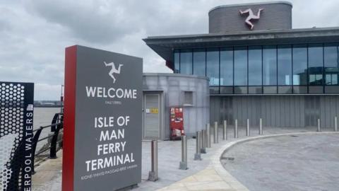 The exterior of the ferry terminal in Liverpool, which has a sign that reads Welcome to the Isle of Man Ferry Terminal. There is a three legs of man sign on the building, which has large windows and is grey. 