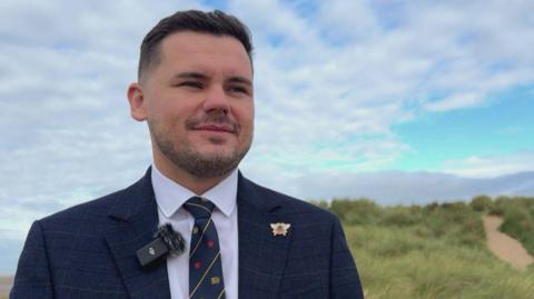 Lancashire County Councillor Joshua Roberts on the sand dunes at Lytham St Annes. The Reform UK politician is wearing a dark blue suit and tie. He has dark hair and is smiling.