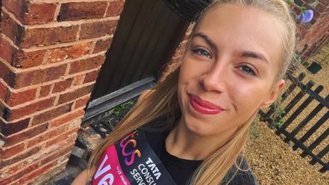 A young woman with blonde hair tied back stands smiling up at the camera for a selfie. She is wearing a black running top.