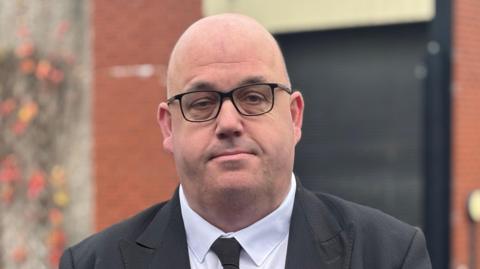 A bald headed man with square black glasses, wearing a black suit, black tie and white shirt.