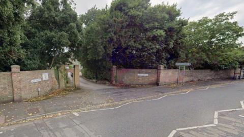 Road with entrance to brick wall lined driveway with dense trees either side.