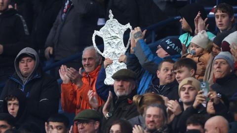 A fan holds a foil FA Cup in the stands