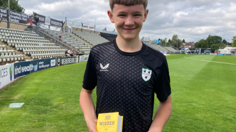 Oliver Whitehouse is in a black cricket shirt and holds a copy of Wisden on a cricket pitch