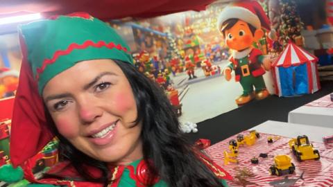 Selfie of Amanda Walker dressed as an elf. She has long black hair and is wearing a red and green bobble hat. She has red make-up on her cheeks and nose. The room behind her is decorated with a Christmas toy theme.