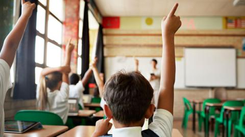 A school classroom with children pictured from behind raising their hands in the air to answer a teacher (stood at front of room in front of whiteboard)
