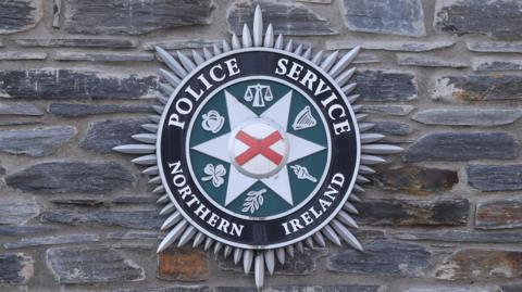 PSNI badge on grey stone wall. The badge is green with symbols on it, a red X in the middle and has a black border with white writing bearing the words .Police Service of Northern Ireland'