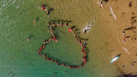 People stood in seawater near the short of a beach. They are wearing red and and are stood in the shape of a heart. The beach is sandy. The water is clear and blue. 