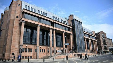 An angled view of Newcastle Crown Court, a large building with dark glass windows. A road runs in front of it.