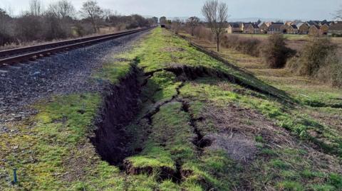 An area of grassland next to a railway line has collapsed. There are trees and a row of houses in the background.