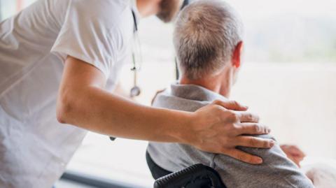 A mature man caregiver with stethoscope is wearing a white top and has his arm around another man in a chair and is leaning down. A man in a chair is looking out the window and you can see the back of his head with short grey hair.