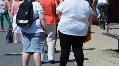 Two people are standing in the street and their T-shirts are tight over their largely-proportioned bodies. You cannot see their faces, only their backs and legs. There are other people around them and a cyclist to their right.