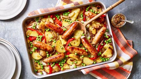 A large tray pan full of sausages, potatoes, peas and chopped red peppers.