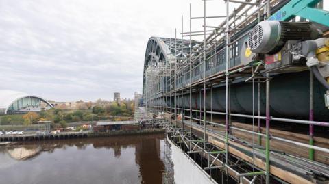 To the right, there is a large green metal arched bridge over the river, with scaffolding on it. In the distance there are trees and buildings and a car park. It is a cloudy day.