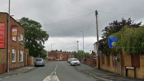 A sign on the right hand side of the road says Baxters Lane. A pub is on the left hand side. Around five cars can be seen parked on either side of the road, with houses seen in the distance.