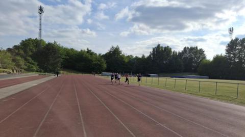 A group of athletes run down a track in the sunshine
