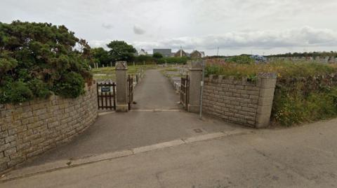 The entrance to the cemetery in St Agnes, Cornwall, on a cloudy day. The entrance has stone walls and two small pillars along with wooden gates and fences. Grass verges with flowers planted in them and some shrubs are also on the border of the site. A number of graves can be seen from the road along with a row of trees and houses in the background.