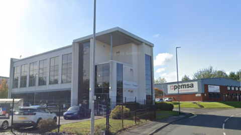 A modern three-storey office building with large glass windows and white exterior cladding stands beside a road on a sunny day. Several cars are parked in front of the building behind a black fence, and a lamppost is visible in the foreground. To the right, a neighbouring commercial building displays the sign “Opemsa Management.” The main office building has signage near the entrance, though the text is not clearly legible. The sky is bright blue with a few small clouds, and landscaped grass and shrubs surround the properties.