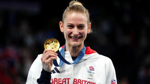 Bryony Page holding her gold medal. She is smiling and wearing a Team GB tracksuit