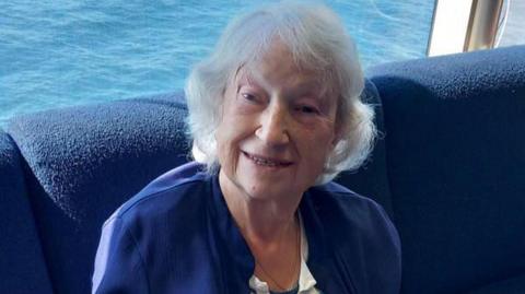 A woman with short white hair layered around her face, smiling while sat inside on a boat. Blue sea can be seen in the background and she is on a cushioned navy seat. She is wearing a navy cardigan and striped white and blue top.