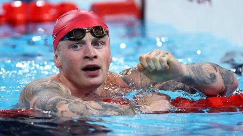 Adam Peaty in the swimming pool with a red swimming cap on. He is leaning on a floating lane divider