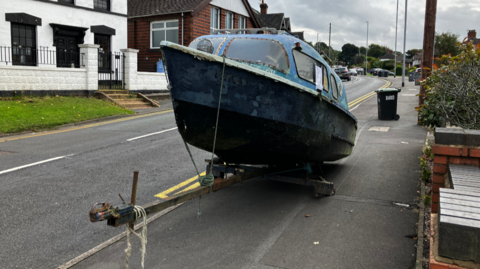 A medium-sized blue boat on a trailer, parked on the edge of a pavement alongside a road.