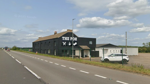 A Google Street View image of the Fox Inn behind green fencing next to the A1 road. It is a large, two-storey black building with a large sign reading "THE FOX" in white capital letters, with three images below - one of a knife and fork, one of a hot tea, and one of a bed. A white Range Rover is parked outside. The sky is blue with white clouds.