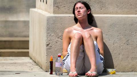 A woman sits on the ground and leaning against a beige stone wall with her knees up near her chest in the blazing sun.  She has a bottle of water on the ground