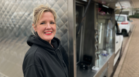 Heidi Hall has blonde hair, and a black hooded top is standing at the back of a silver coloured catering vehicle. She is smiling at the camera