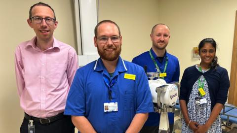 members of the team stood in a row. all of them are looking directly at the camera and smiling. Marcus Dakers and Stuart McGrail are wearing scrubs.